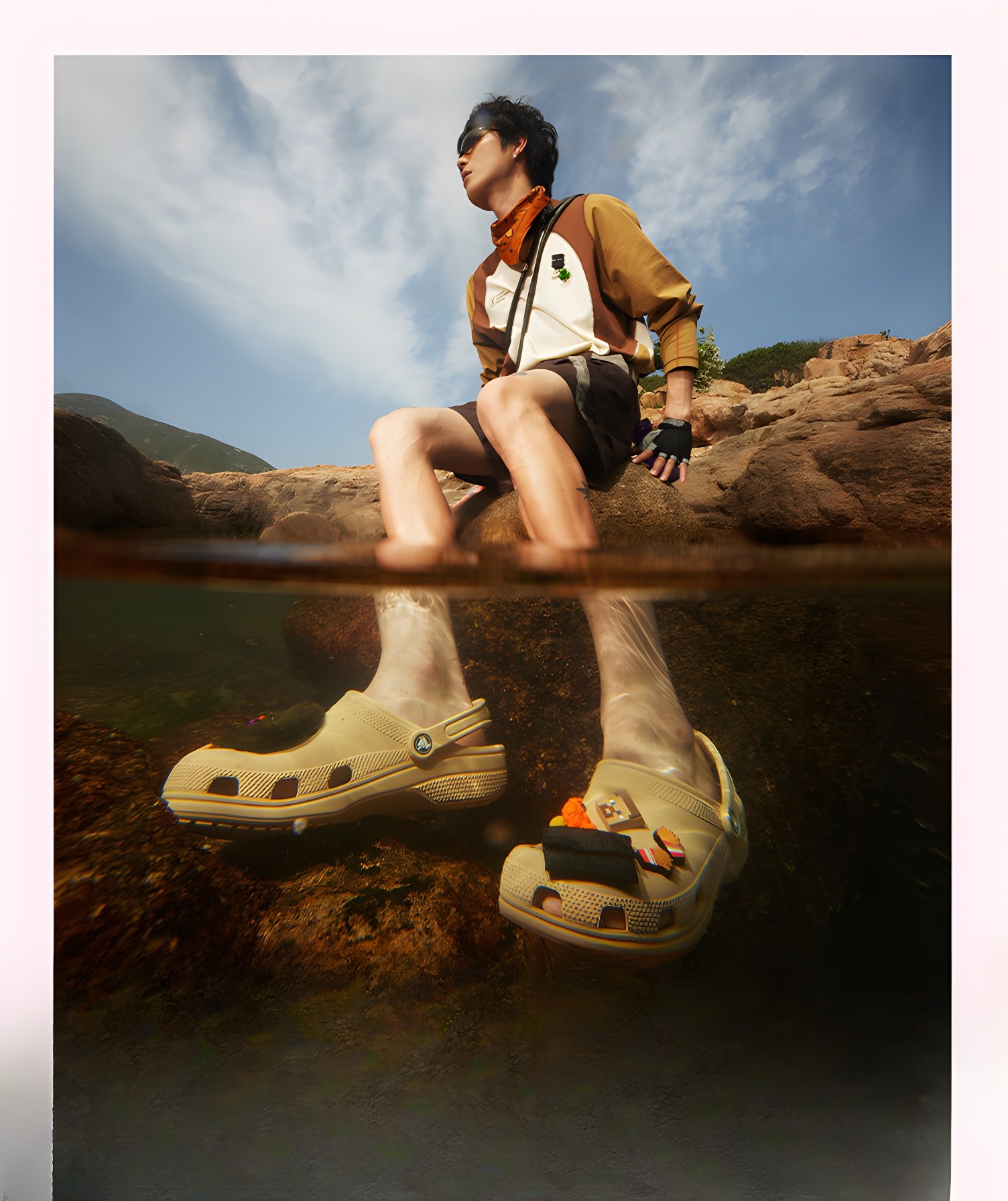 A man sitting on rocks by the water, wearing beige CROCS clogs with Jibbitz charms, captured in a split-level shot showing both above and below the waterline